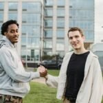 Young diverse male friends shaking hands while standing on lawn against modern building and looking at camera
