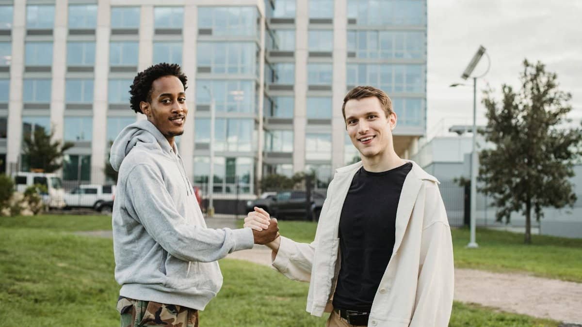 Young diverse male friends shaking hands while standing on lawn against modern building and looking at camera