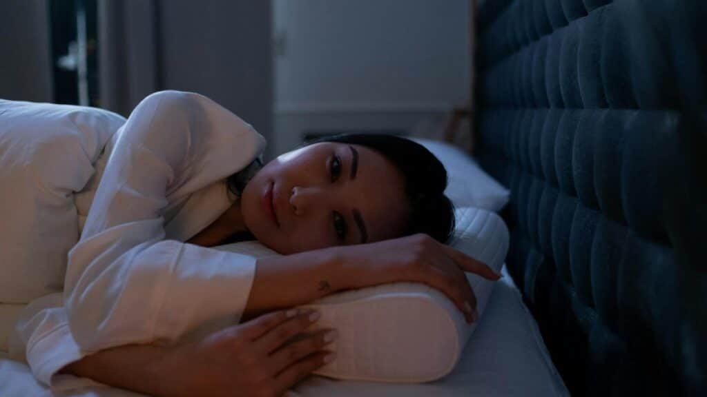 Dark bedroom scene of an Asian woman with insomnia lying on her side, looking thoughtful.