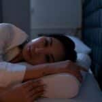 Dark bedroom scene of an Asian woman with insomnia lying on her side, looking thoughtful.