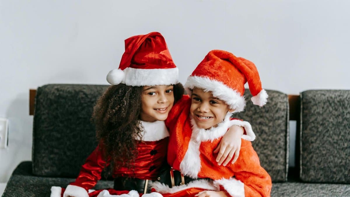 Content black siblings in Santa costumes embracing on couch during New Year holiday and looking away in house