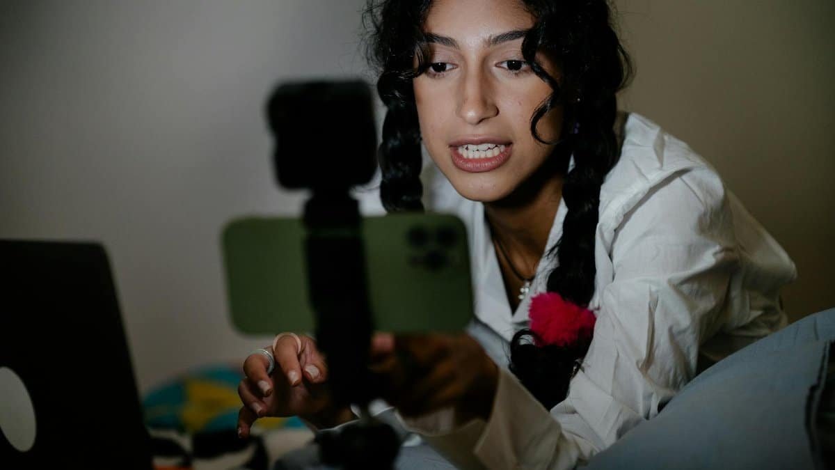 A young woman with braided hair records a video using her smartphone and laptop indoors.