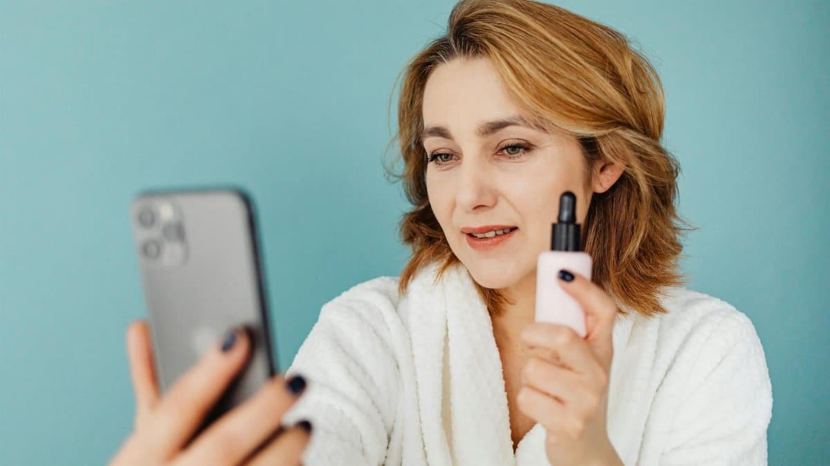 Woman in a white robe taking a selfie while holding a cosmetic bottle against a blue background.