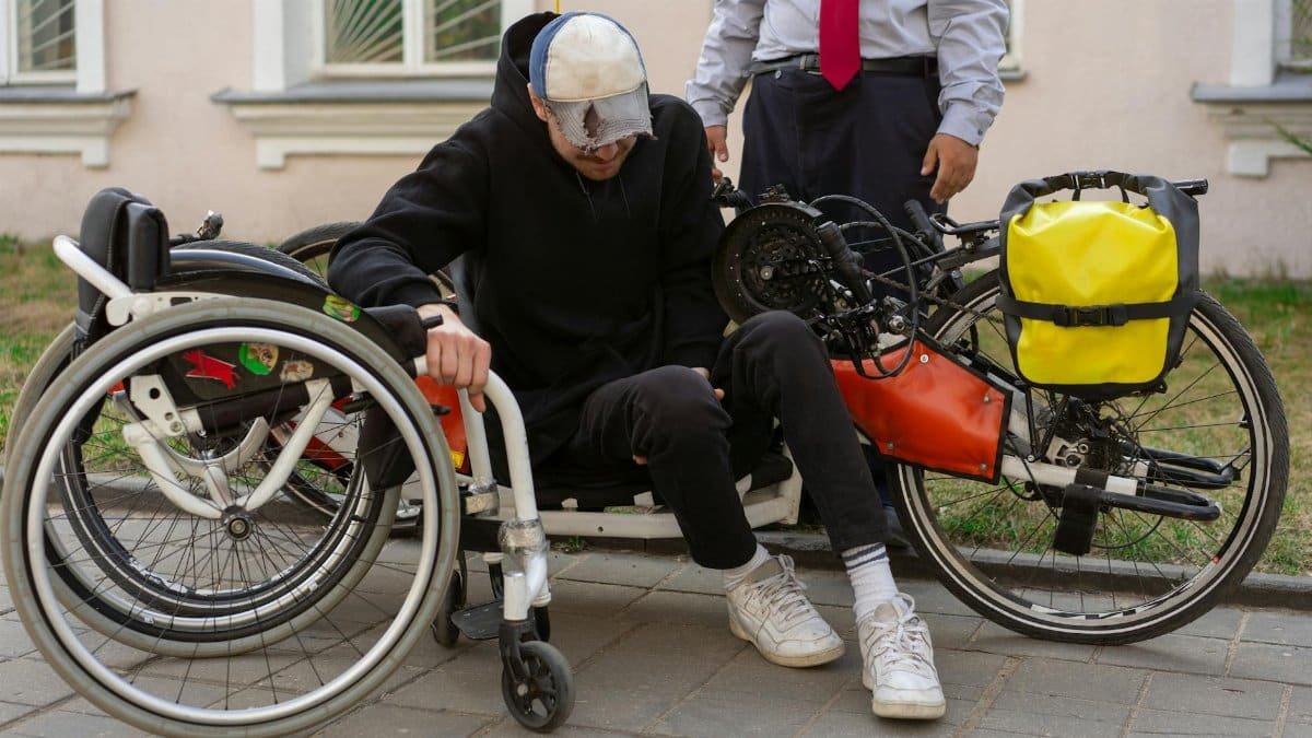 A man transferring from a wheelchair to a handcycle on an urban sidewalk, assisted by another person.