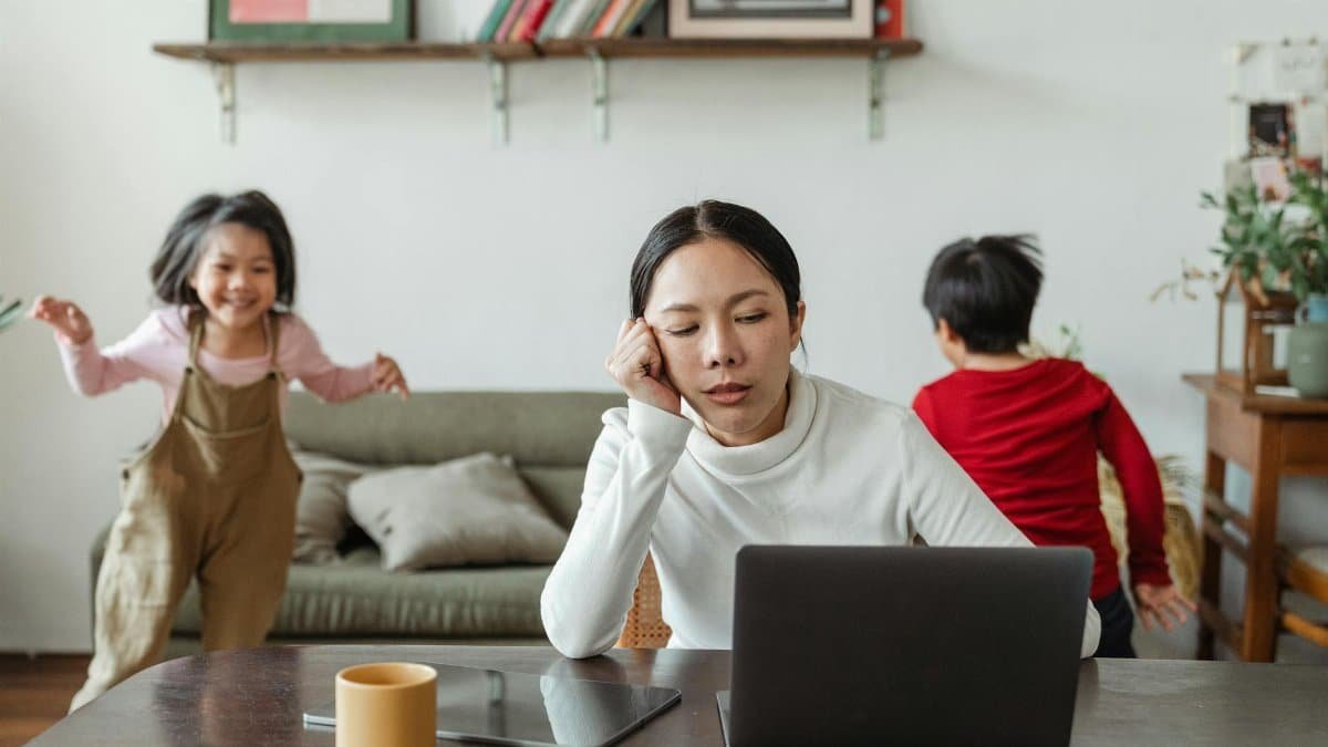 A tired mother working on a laptop while her children play around indoors, highlighting remote work challenges.