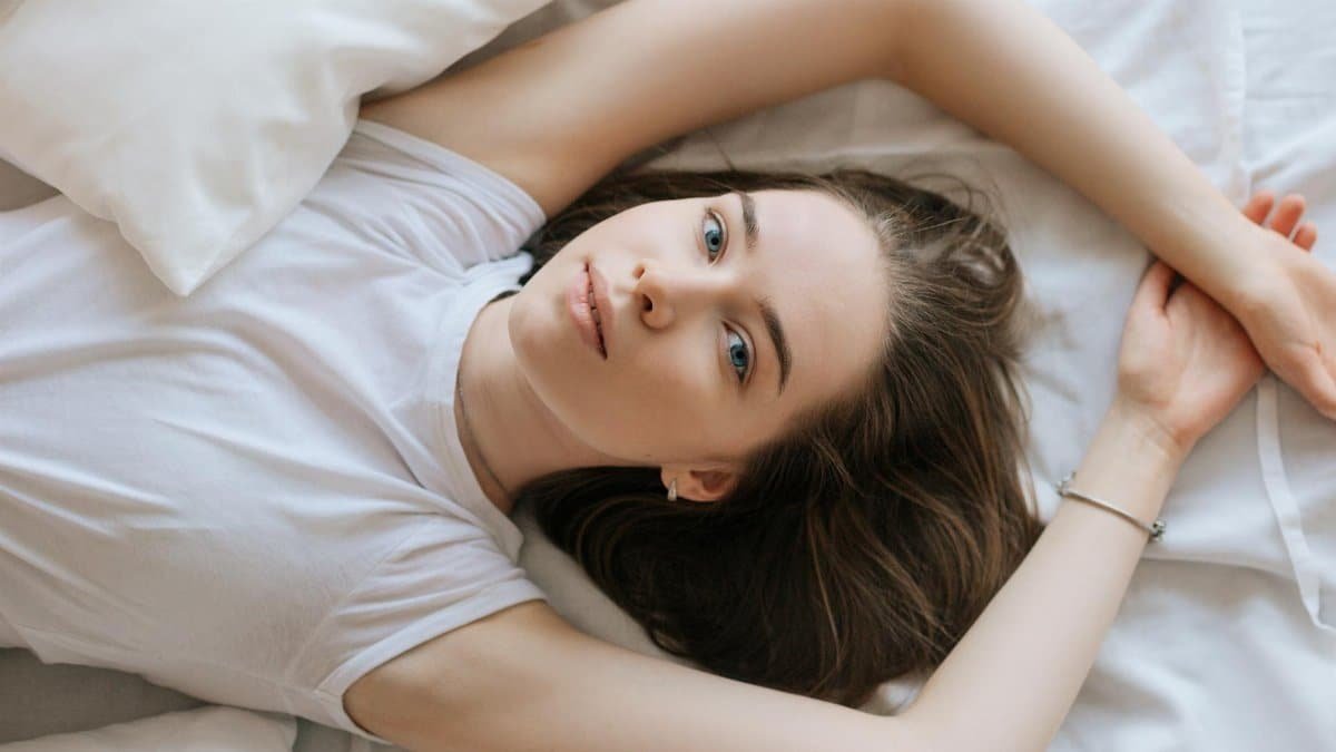 A woman lying down on her bed with white linens, in a relaxed pose wearing a white shirt.