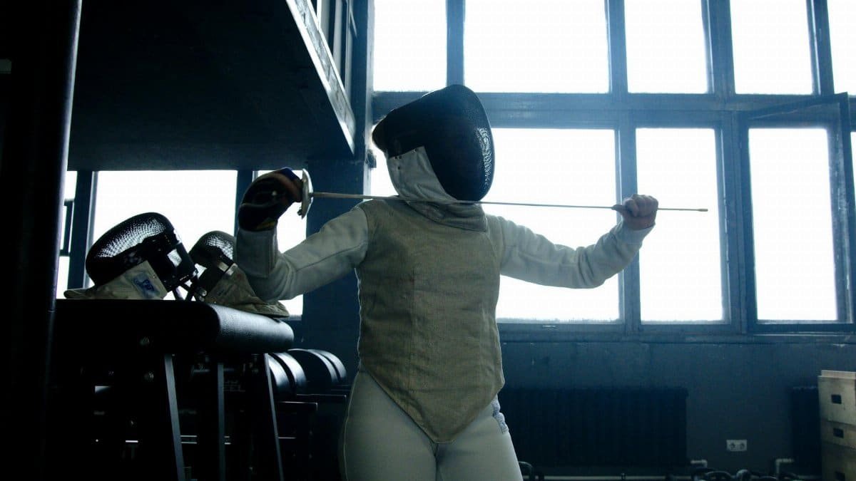 Female fencer in full gear practices her sword technique indoors under natural light.