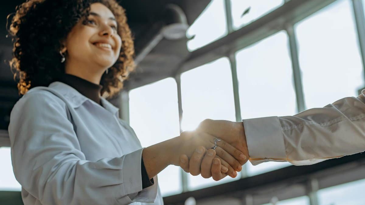 A smiling woman with curly hair shaking hands in a bright, modern office environment.