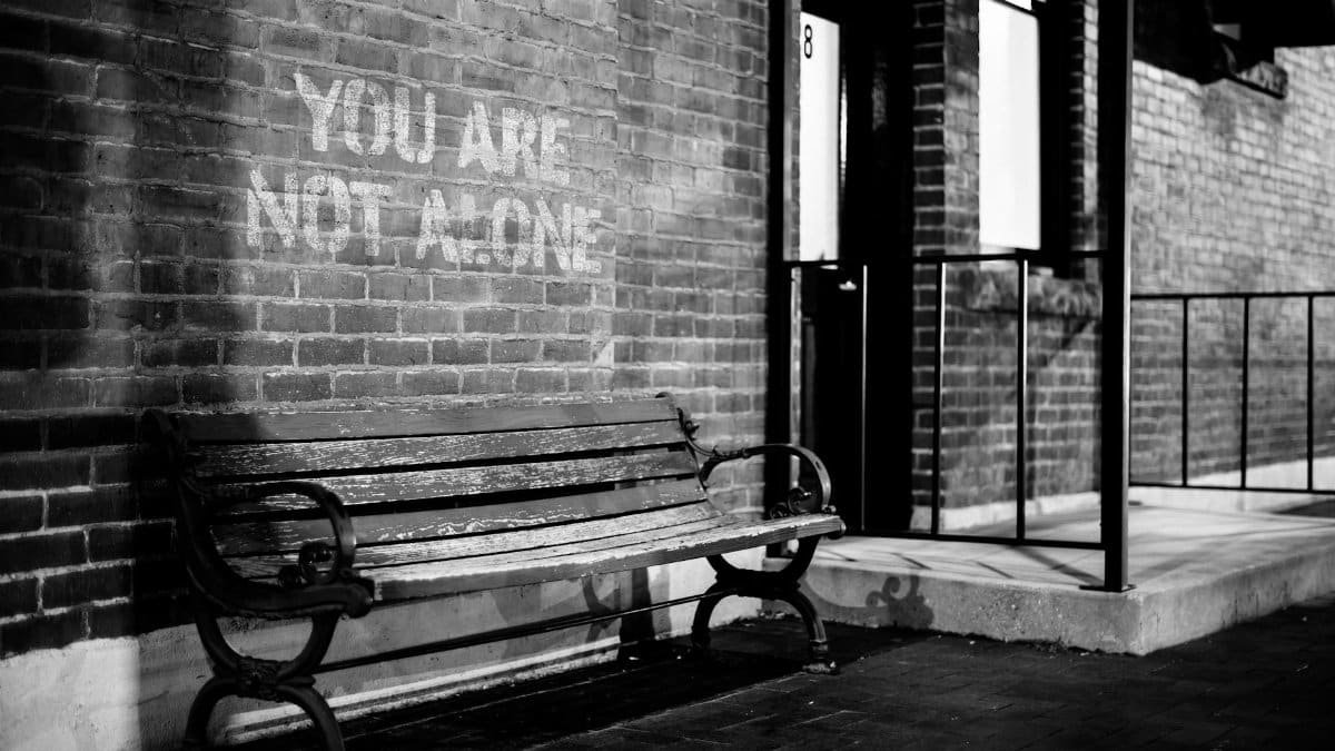 Black and white of empty wooden bench near brick wall of building with inscription you are not alone
