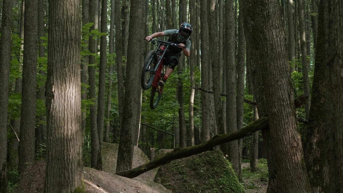 Mountain biker jumps over a trail in a dense forest, showcasing extreme sports skills.