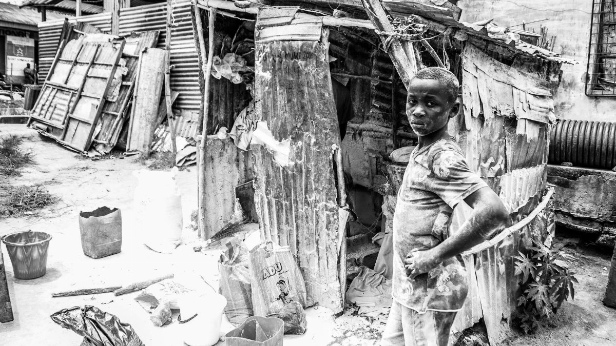 Black and white photo of a young boy standing in front of a makeshift structure in Kinshasa, depicting urban life and resilience.