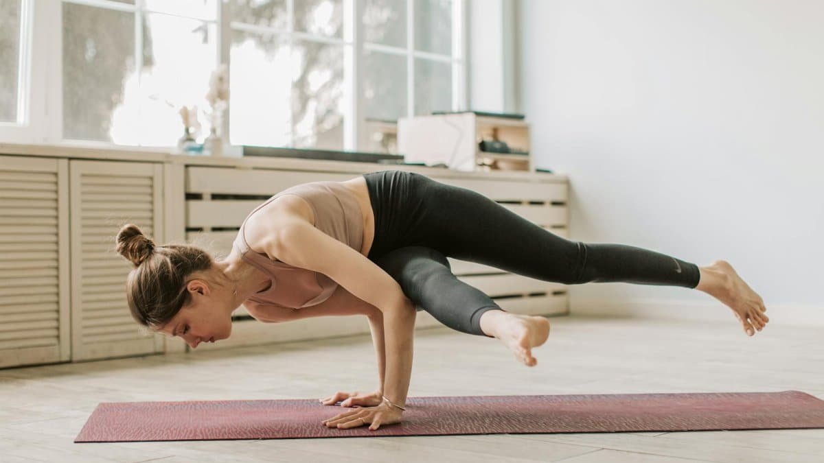 A woman performs an advanced yoga pose on a mat in a bright, modern indoor setting.