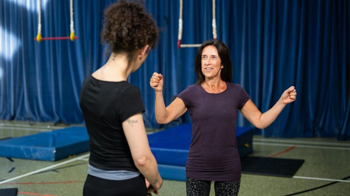 Two women engaged in a trapeze class inside a gym with mats and ropes.