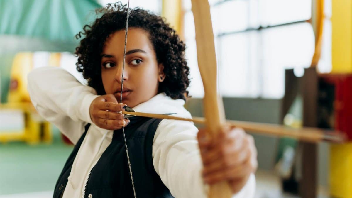 Young woman with a determined expression practicing archery in an indoor setting.