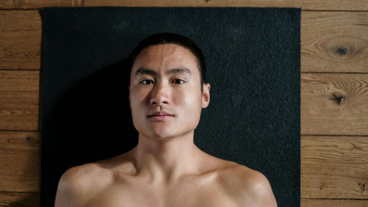 A shirtless man relaxing on a black yoga mat in a wooden floored studio.