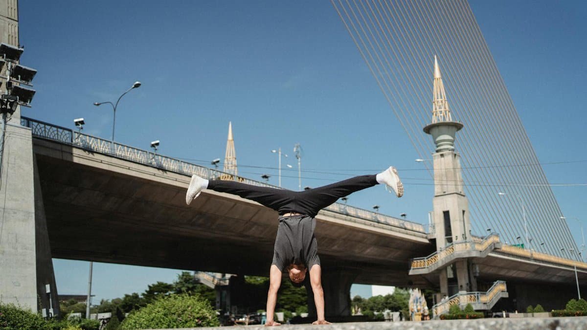 Full length strong flexible sportsman in dark activewear doing handstand and split in air while exercising on street on elevated highway background