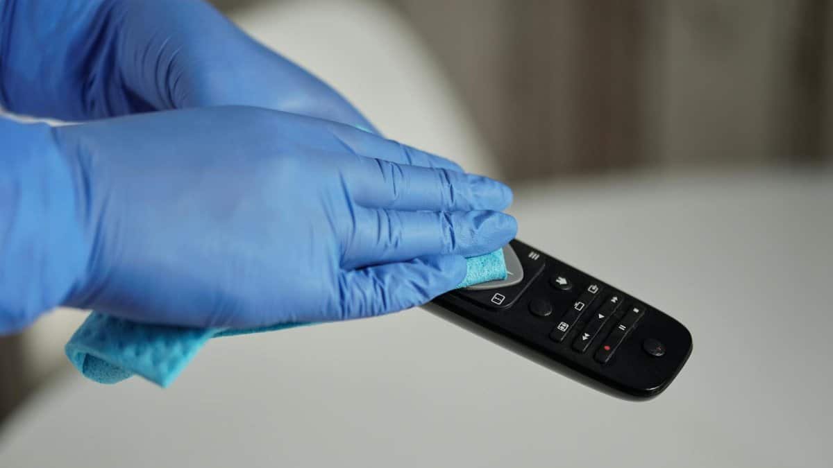 Hands in gloves sanitizing a remote control for hygiene purposes, emphasizing cleanliness.
