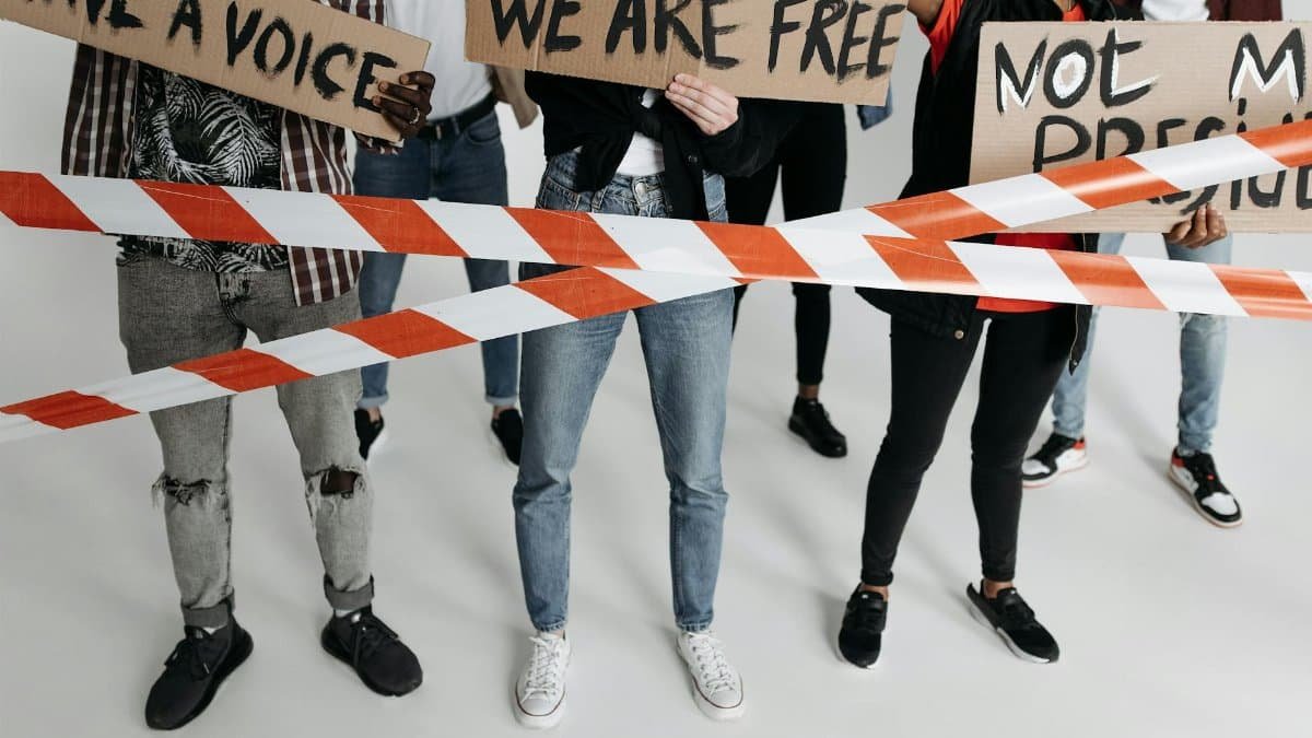 A diverse group of adults holding protest signs behind tape in a studio setting.