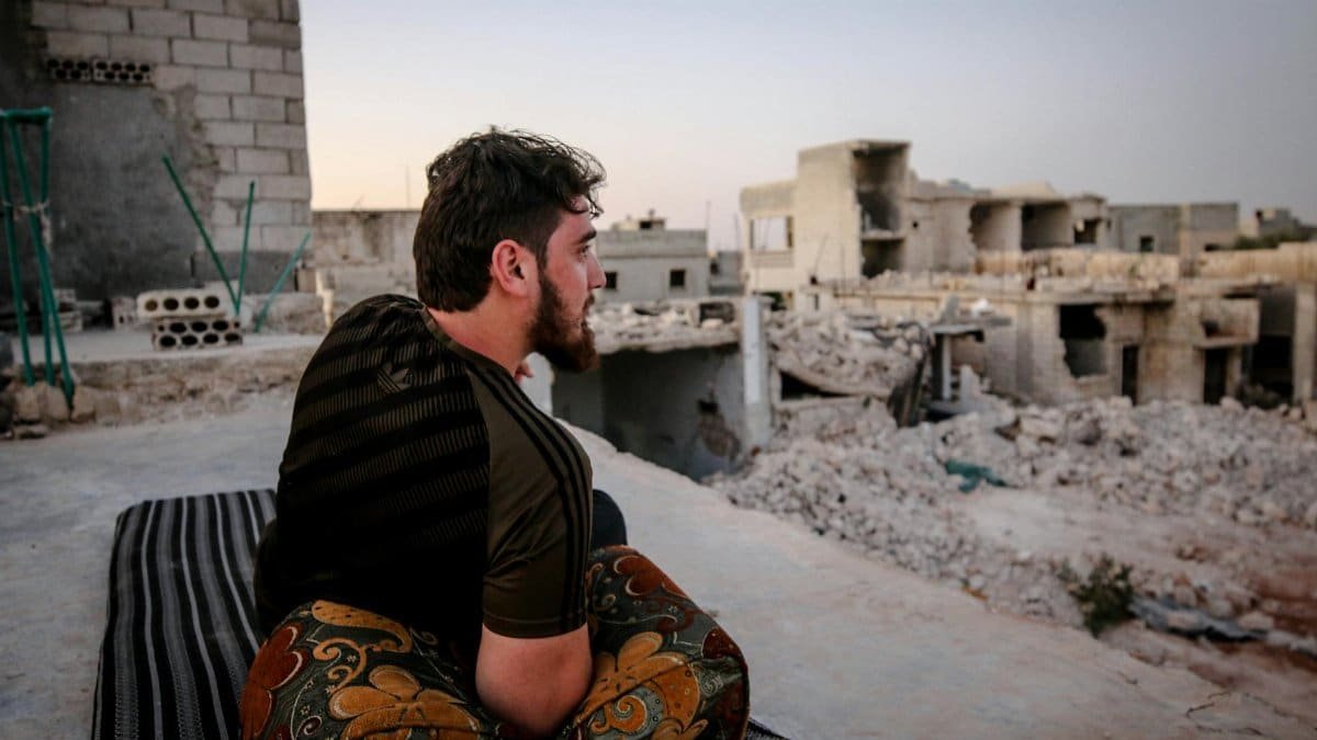 A man in Idlib, Syria, sits thoughtfully amidst the ruins of buildings, reflecting the impact of conflict.