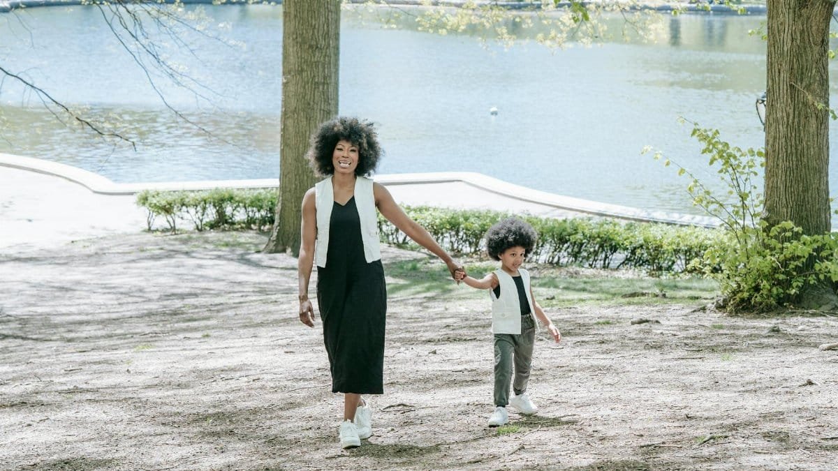 A mother and her child walk hand in hand by a peaceful lakeside in a sunny park.