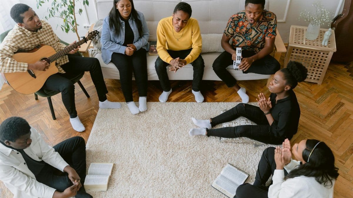 A diverse group of people praying, singing, and playing guitar indoors, emphasizing faith and community.