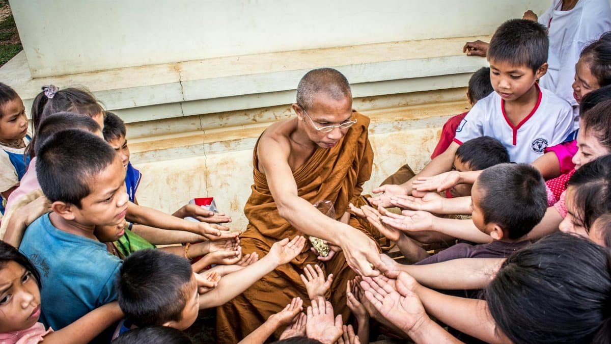 A monk shares offerings with a group of children outdoors, promoting compassion and connection.