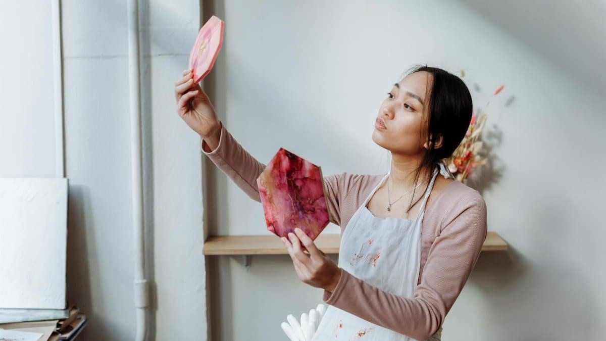 An Asian woman artist in a studio holds and examines vibrant resin art pieces with focus.