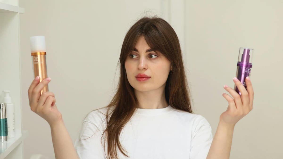 A young woman compares two cosmetic products while standing indoors, unsure of which to choose.