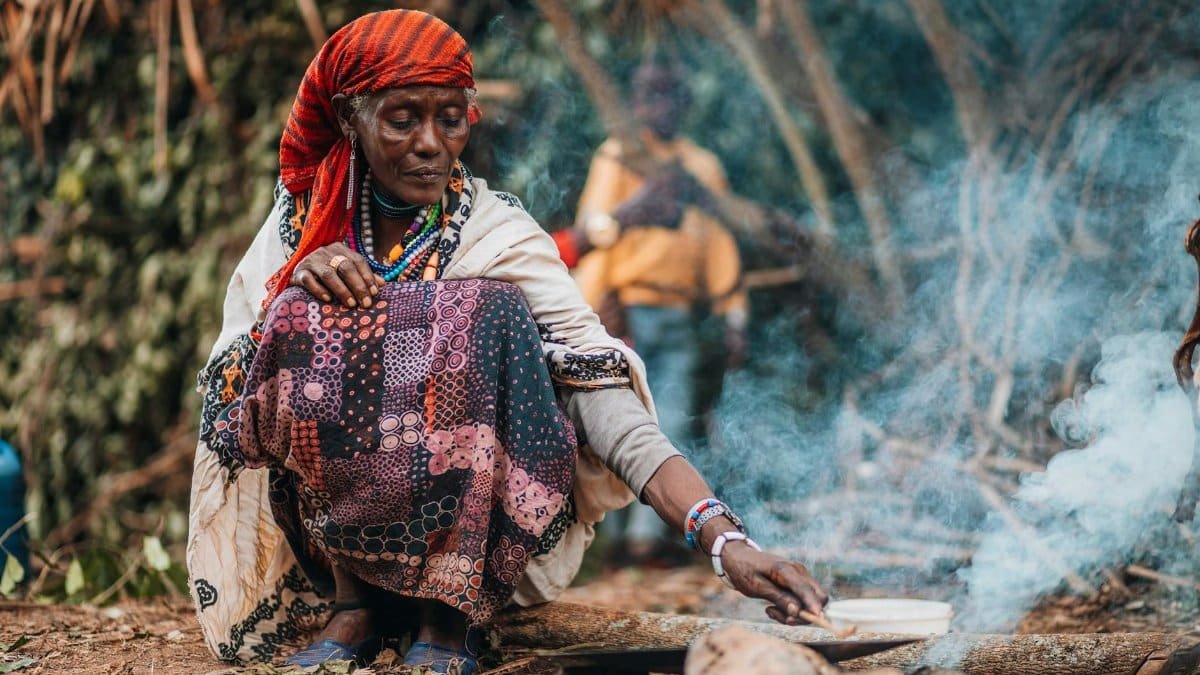 A woman cooking outdoors in a traditional African village setting with visible smoke.