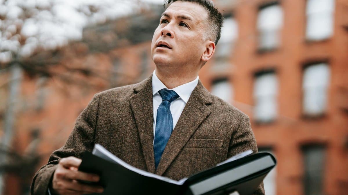 A professional man in a formal coat looks upwards while holding a folder outdoors.