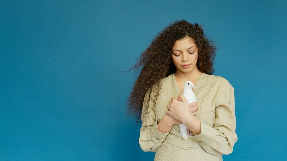 Portrait of a young woman in a beige dress holding a white dove against a blue backdrop.