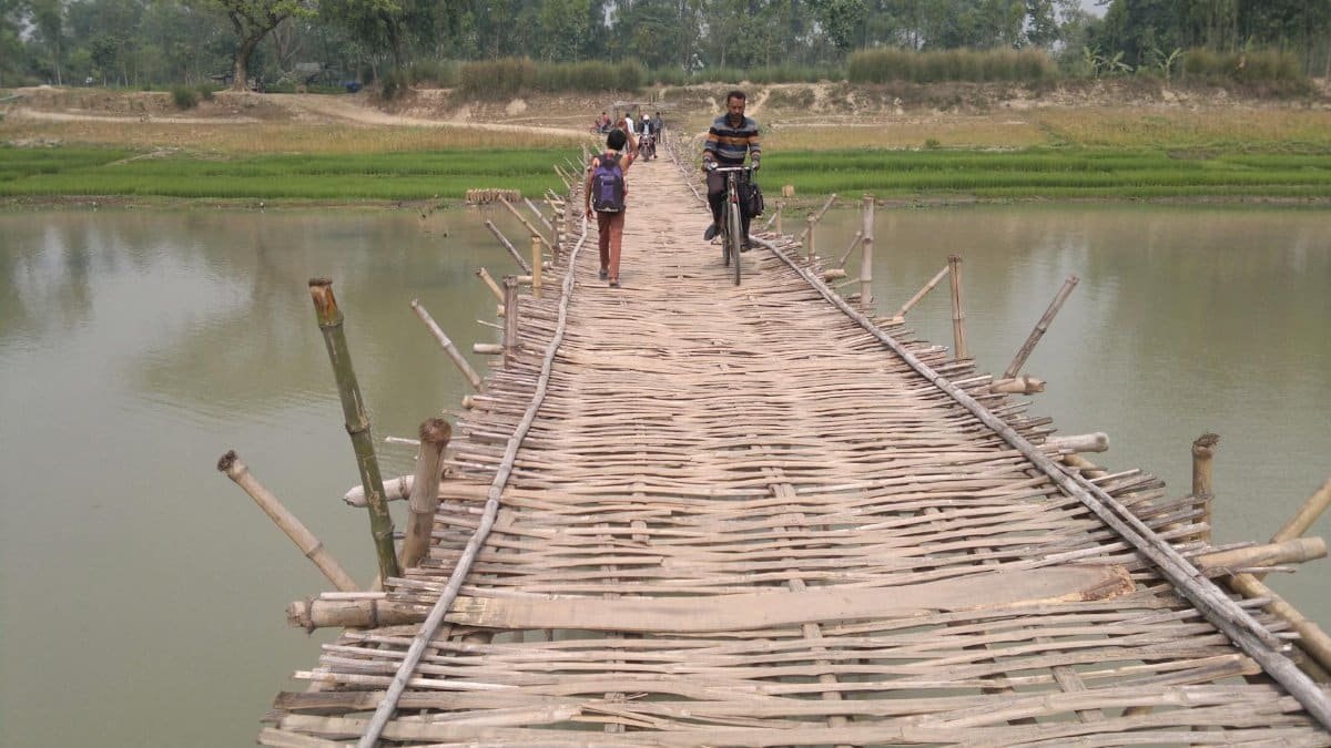 A rustic bamboo footbridge crossing a calm river with people walking and a man bicycling.