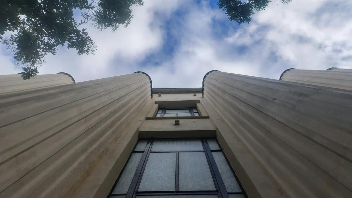 Low-angle shot of a beige modern building with a sky background framed by trees, capturing an upward architectural perspective.