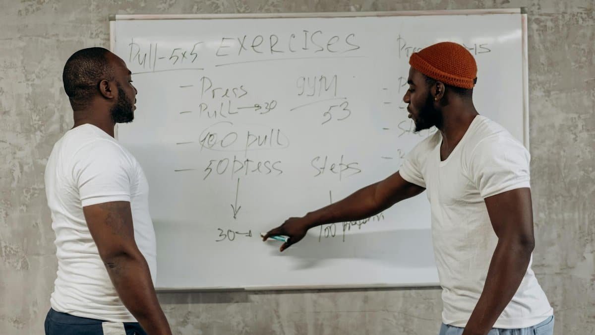 Two men discussing exercise routines on a whiteboard, planning gym workouts indoors.