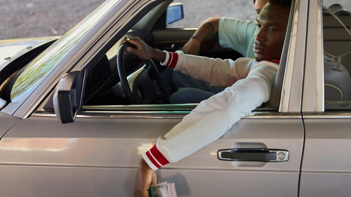 A young adult man sits in a vintage car, casually holding a wad of cash through the window.