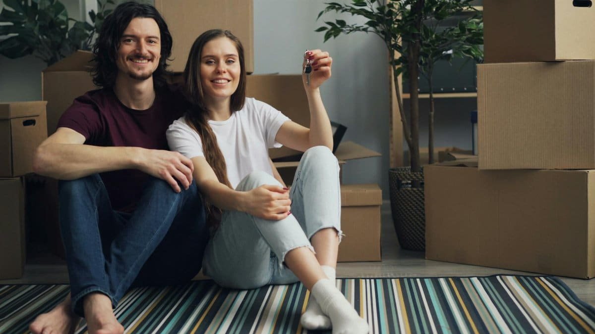 A joyful couple sitting on a striped carpet with moving boxes, holding a key to their new home.