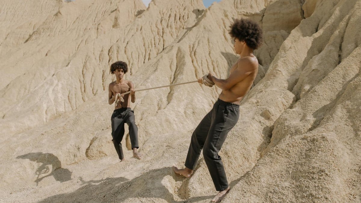 Two shirtless men with afro hair participate in a tug of war challenge in a rocky desert.
