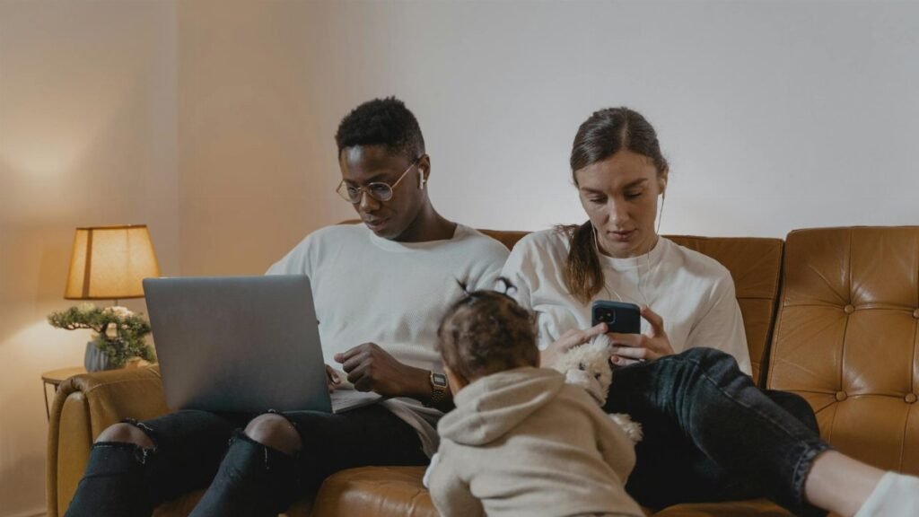 A family sitting on a couch using a laptop and phone while a toddler plays nearby. Cozy indoor setting.