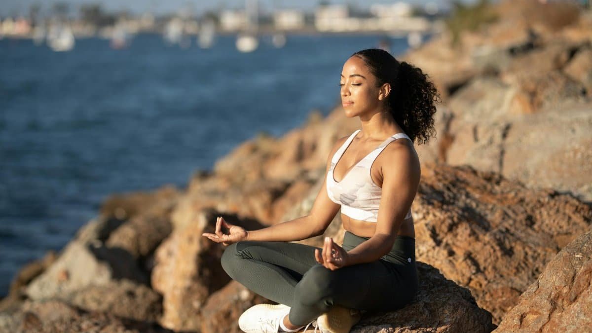 Woman meditating on rocks by the ocean, embodying calmness and wellness.
