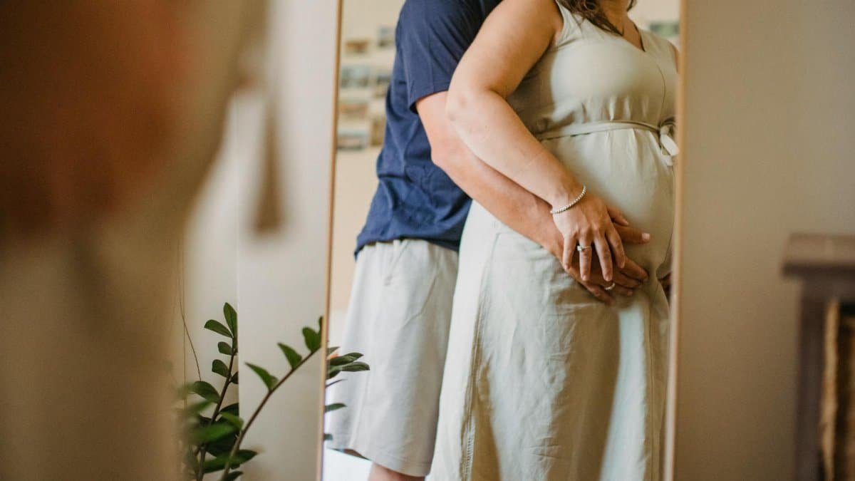 A loving couple embraces while expecting, captured in a warm indoor setting with a gentle tone.