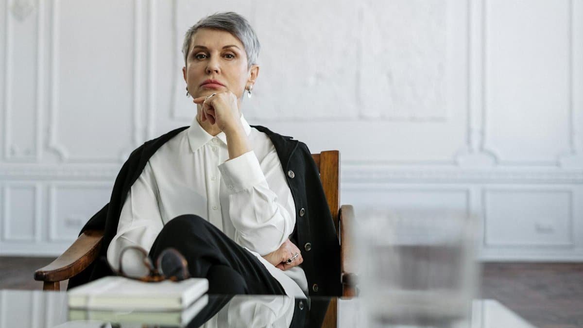 A thoughtful professional woman sits at a glass table in a well-designed office, conveying contemplation and expertise.