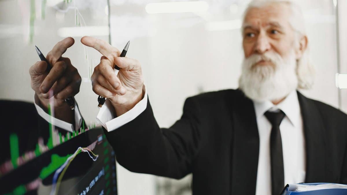 Elderly businessman with beard presenting financial trends on a screen.