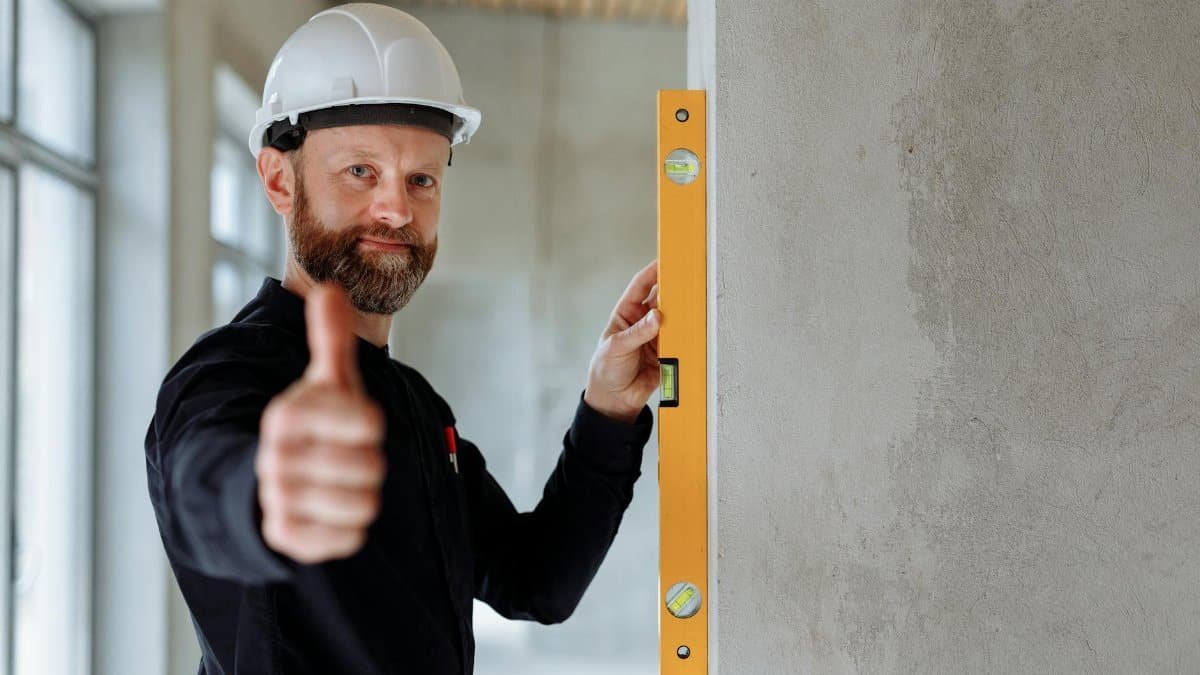 Professional construction worker with hard hat and spirit level giving thumbs up indoors.