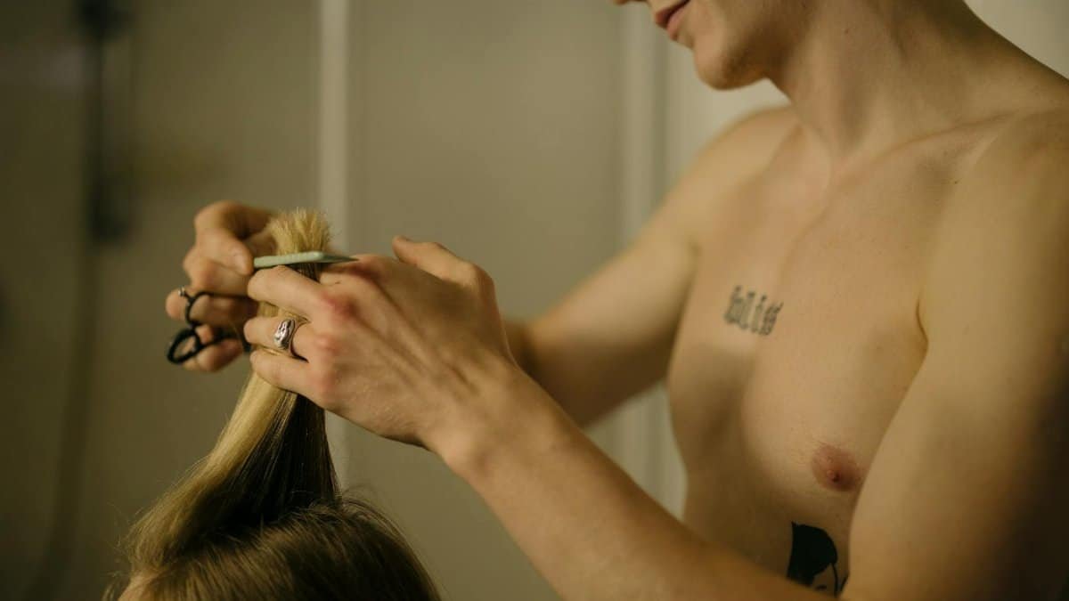 A shirtless tattooed barber skillfully cutting blonde hair indoors, showcasing haircut skills.