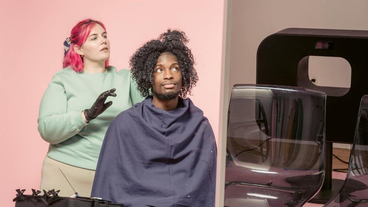 Skilled female hairdresser touching Afro hair of African American male client while sitting near professional supply on pink background in studio