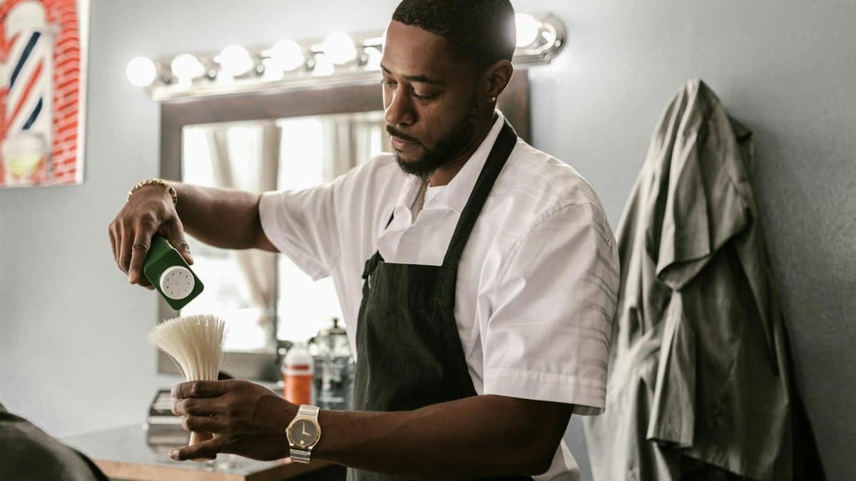 A barber in an apron prepares a brush inside an indoor barber shop, focusing on grooming tools.