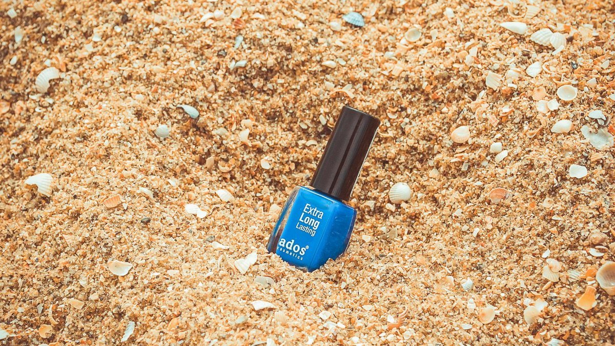 Vibrant blue nail polish bottle nestled in sandy beach setting with seashells.