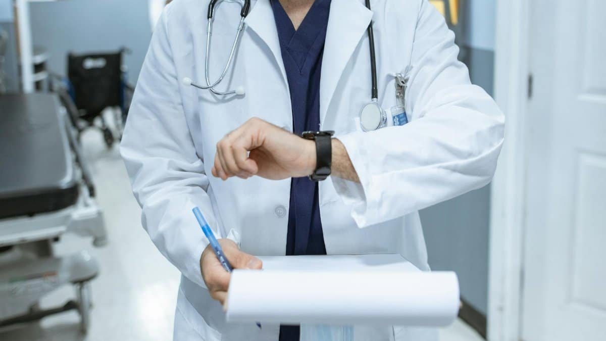 Doctor in lab coat checking wristwatch while holding medical chart in hospital corridor.