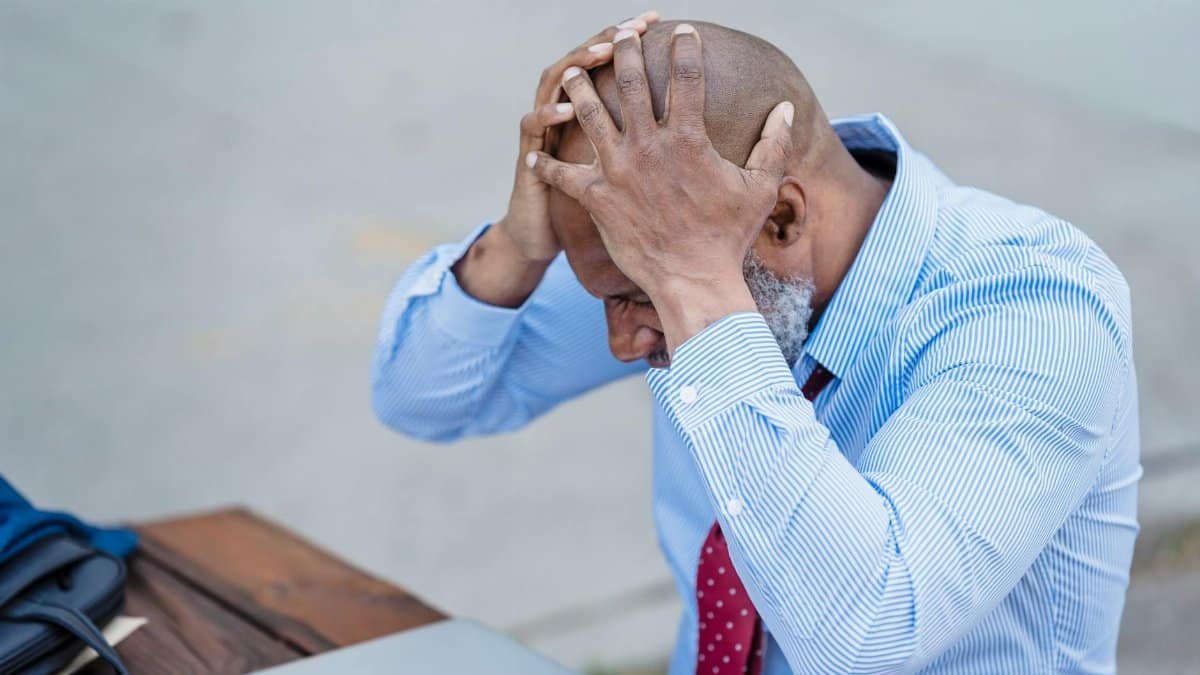 Frustrated businessman holding head in hands outdoors with laptop, corporate stress concept.
