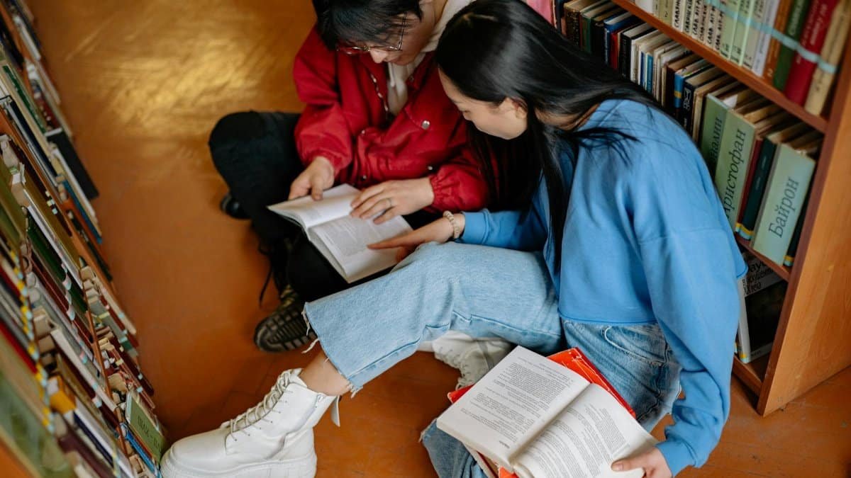 Two students sitting and reading books in a library aisle, exploring literature.